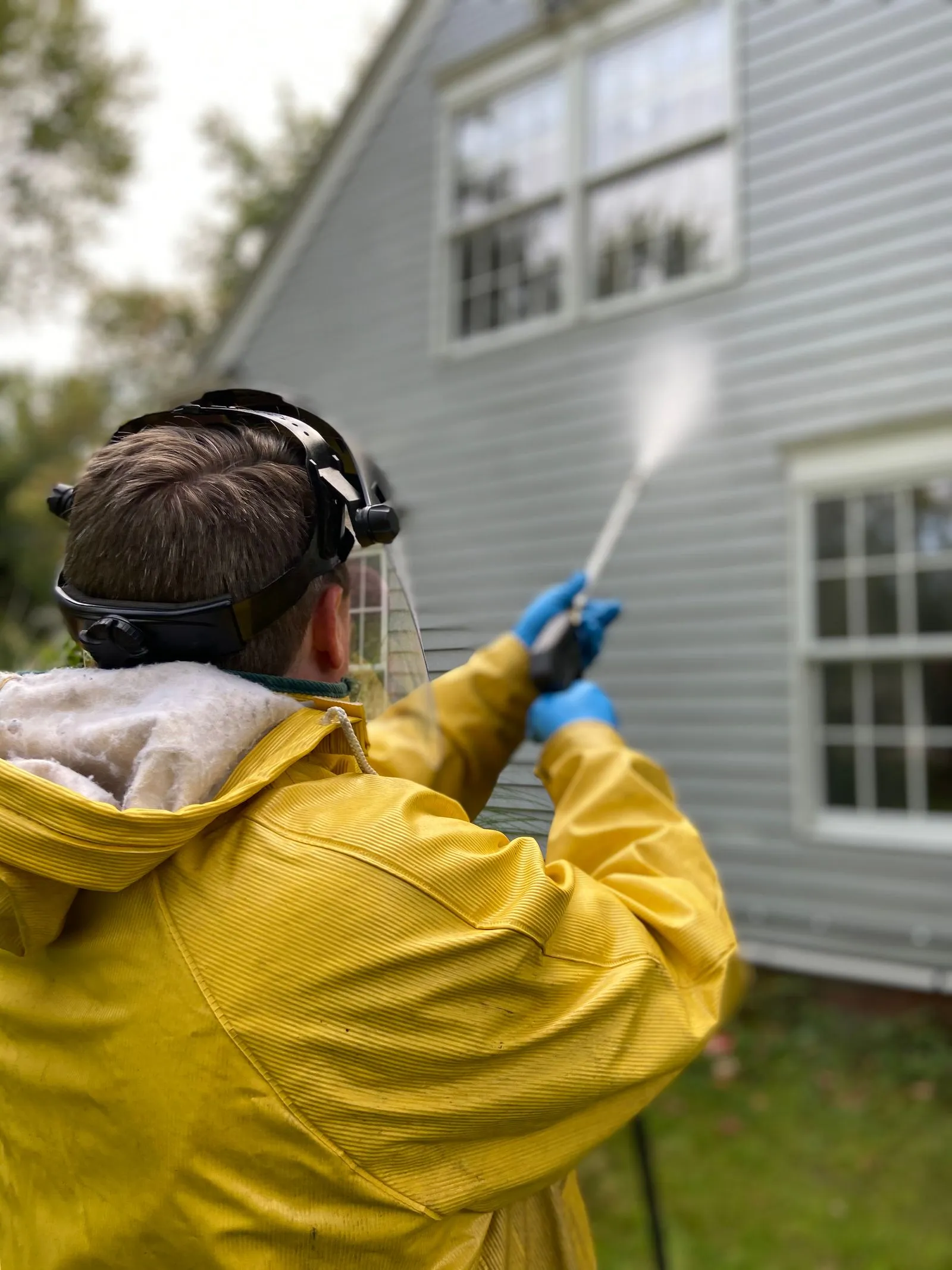 Technician in rain gear soft washing the siding of a two-story house